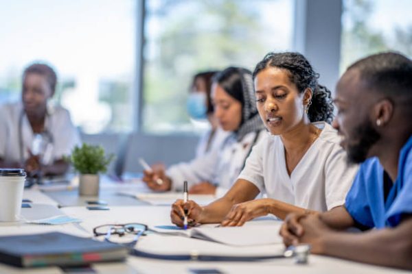 istockphoto-1399300572-612×612 A small group of medical professionals sit around a boardroom table as they meet to discuss patient cases.  They are each dressed professionally in scrubs and lab coats as they work through the case files spread out on the desk collaboratively in small groups.